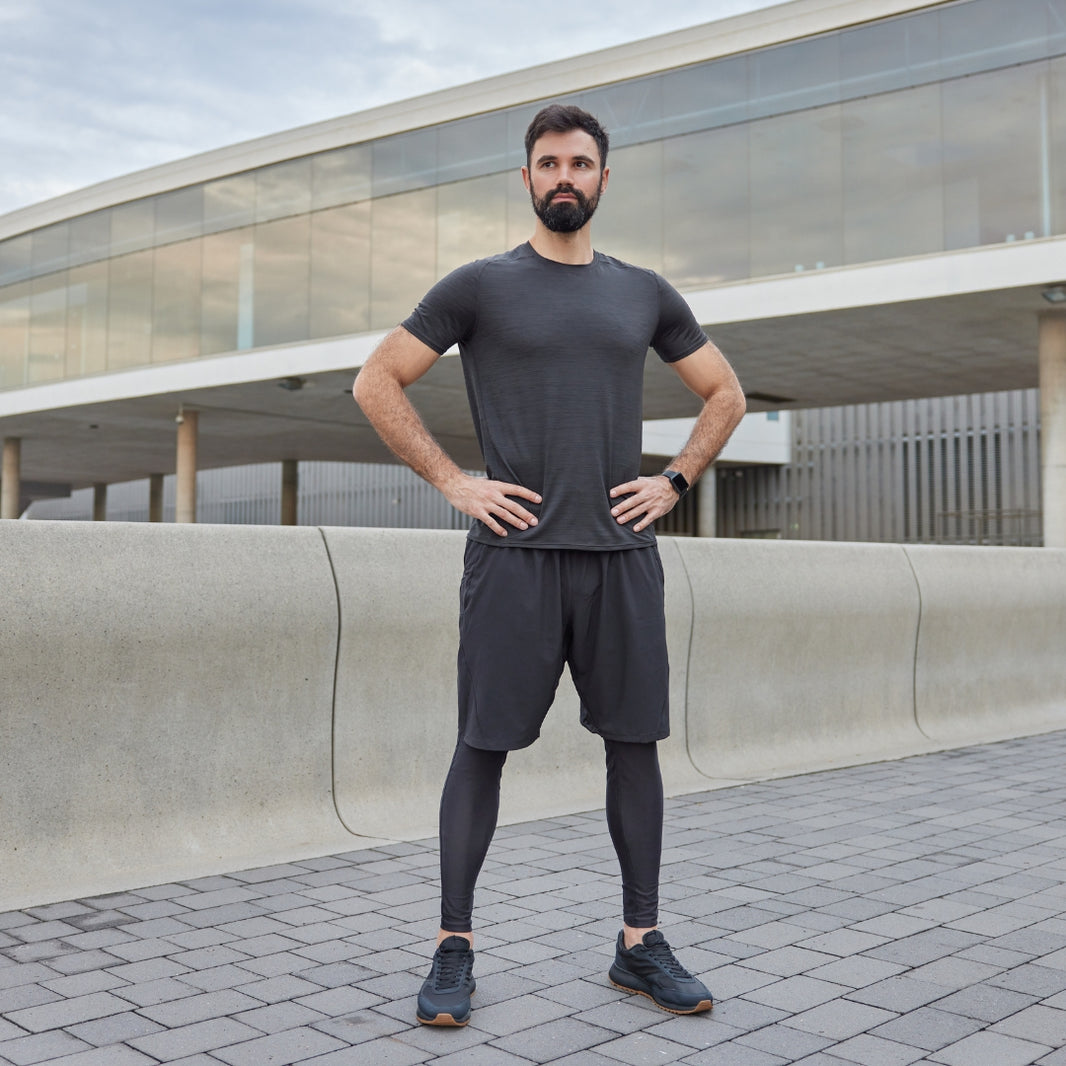 A man wearing a dark gray athletic t-shirt, matching shorts, and compression leggings stands with hands on hips in an outdoor urban setting with a modern glass building in the background.