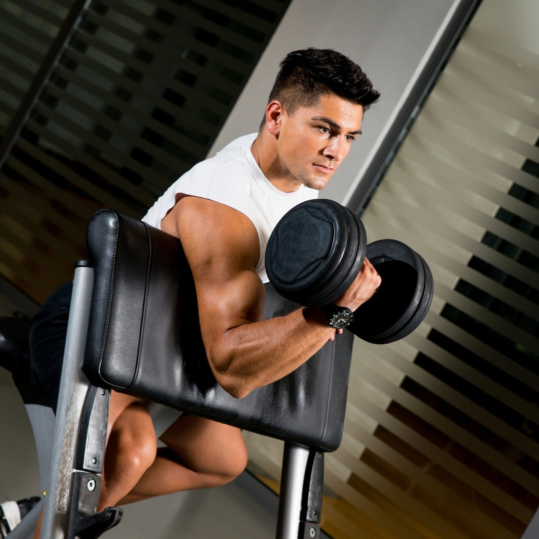 A muscular man in a white sleeveless shirt performs a dumbbell shoulder press on a black padded gym bench, holding a black dumbbell in each hand.