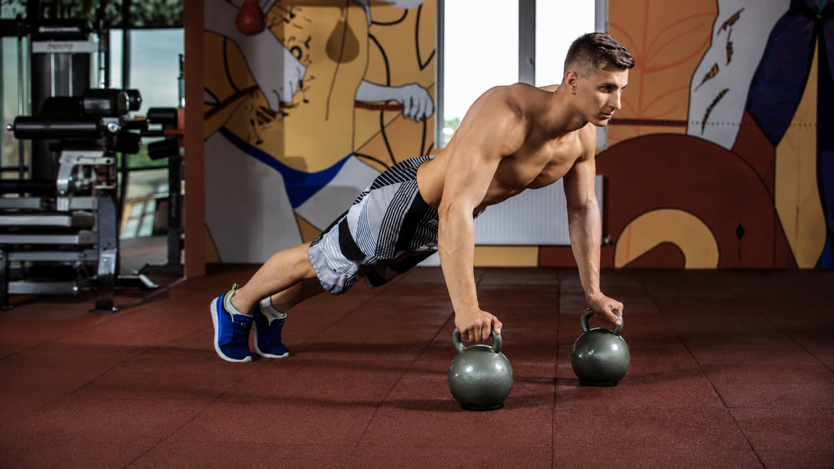 A shirtless man in a gym performing a push-up position with two kettlebells on the floor, wearing black and white patterned shorts and blue sneakers.
