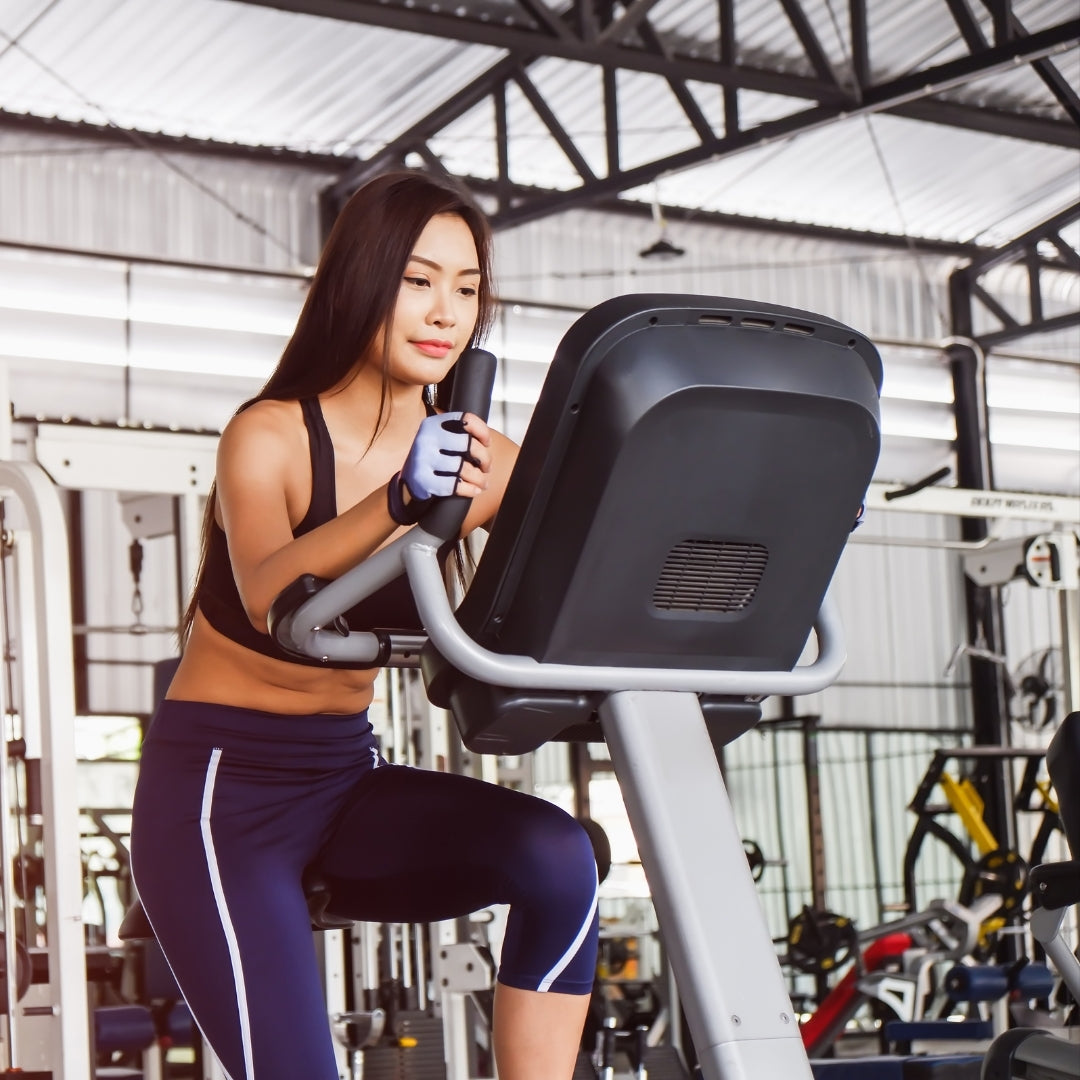 A woman in a black sports bra and navy athletic capri leggings with white side stripes is using a grey and black elliptical machine inside a gym.