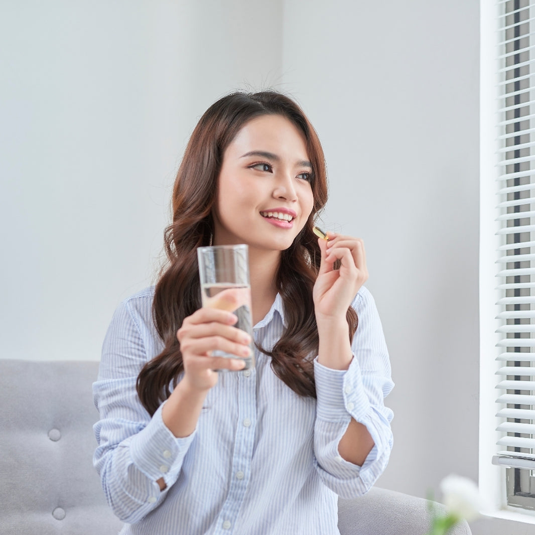 A woman with long brown hair sits on a gray tufted sofa, wearing a light blue and white striped button-up shirt. She holds a clear glass of water in one hand and a small yellow snack in the other, looking to the side with a smile.
