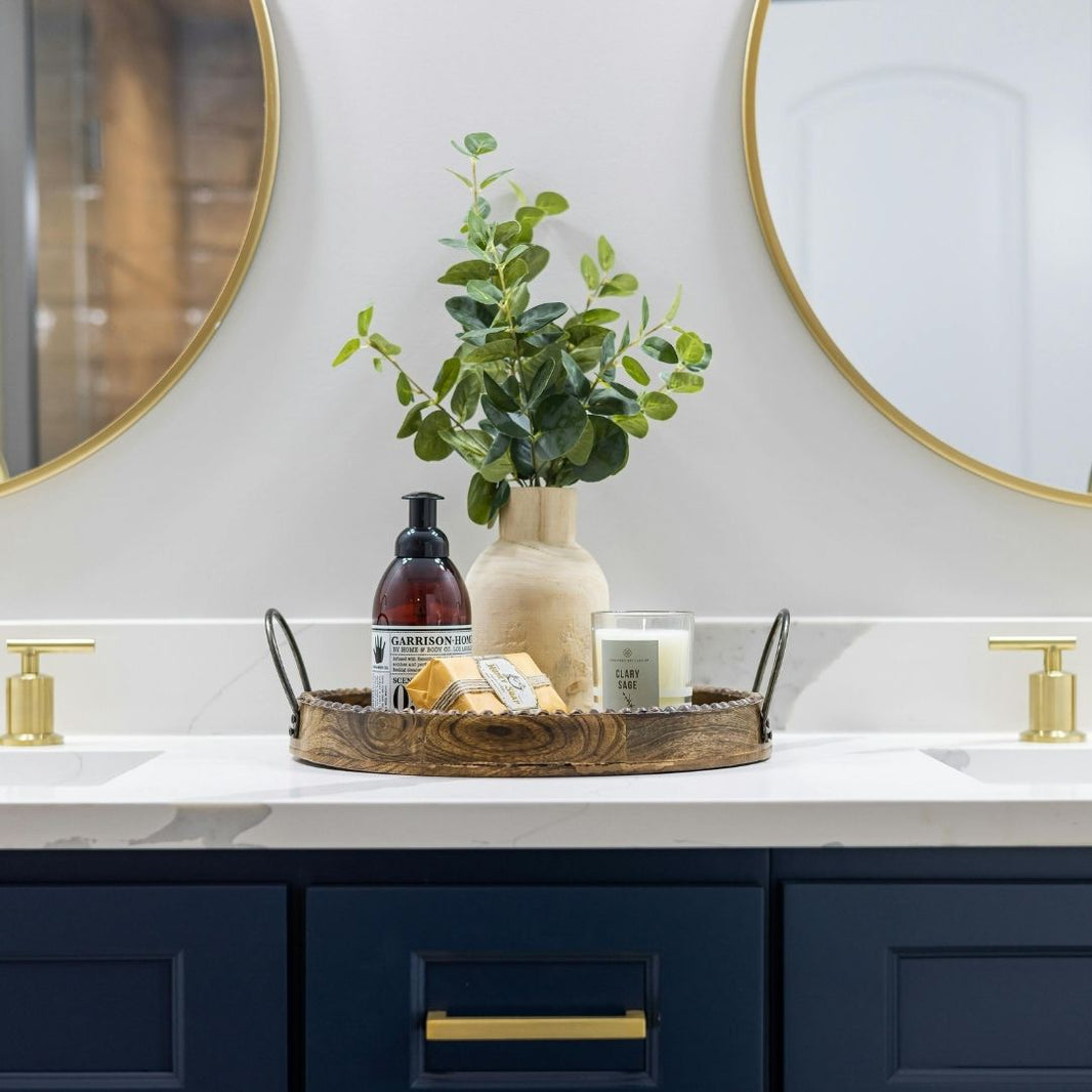 A round wooden serving tray with black metal handles sits on a white marble bathroom countertop, styled with a beige ceramic vase of green leaves, a dark amber bottle, a candle, and soap bars. The tray is positioned in front of a white wall with two round gold-framed mirrors and a navy blue vanity with gold hardware.